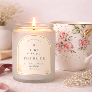 Candle with 'Here Comes the Bride' message in a decorative jar on a table with flowers and teacup.