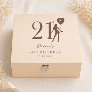 A square light wood keepsake box with a front clasp, engraved with '21st Birthday' and a silhouette figure holding a balloon, styled on a cream fabric background with dried flowers and a ribbon.
