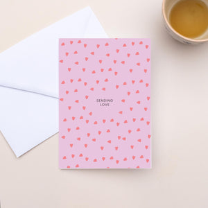 Pink card with heart pattern and white envelope on a beige surface