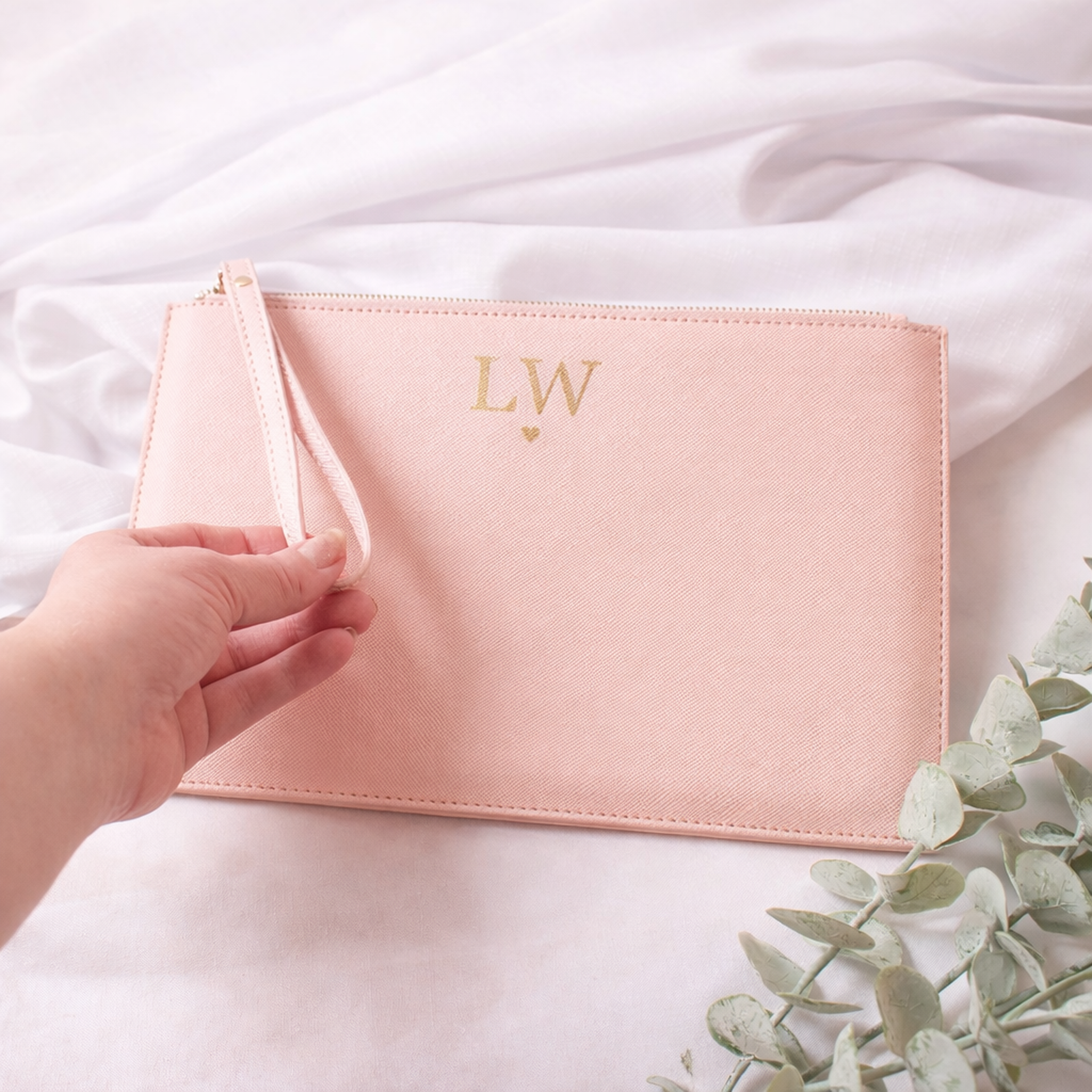 Pink pouch with initials 'LW' held by a hand on a white fabric background with green leaves.