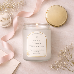 Candle in a jar labeled 'Here Comes the Bride' on a light surface with decorative elements.