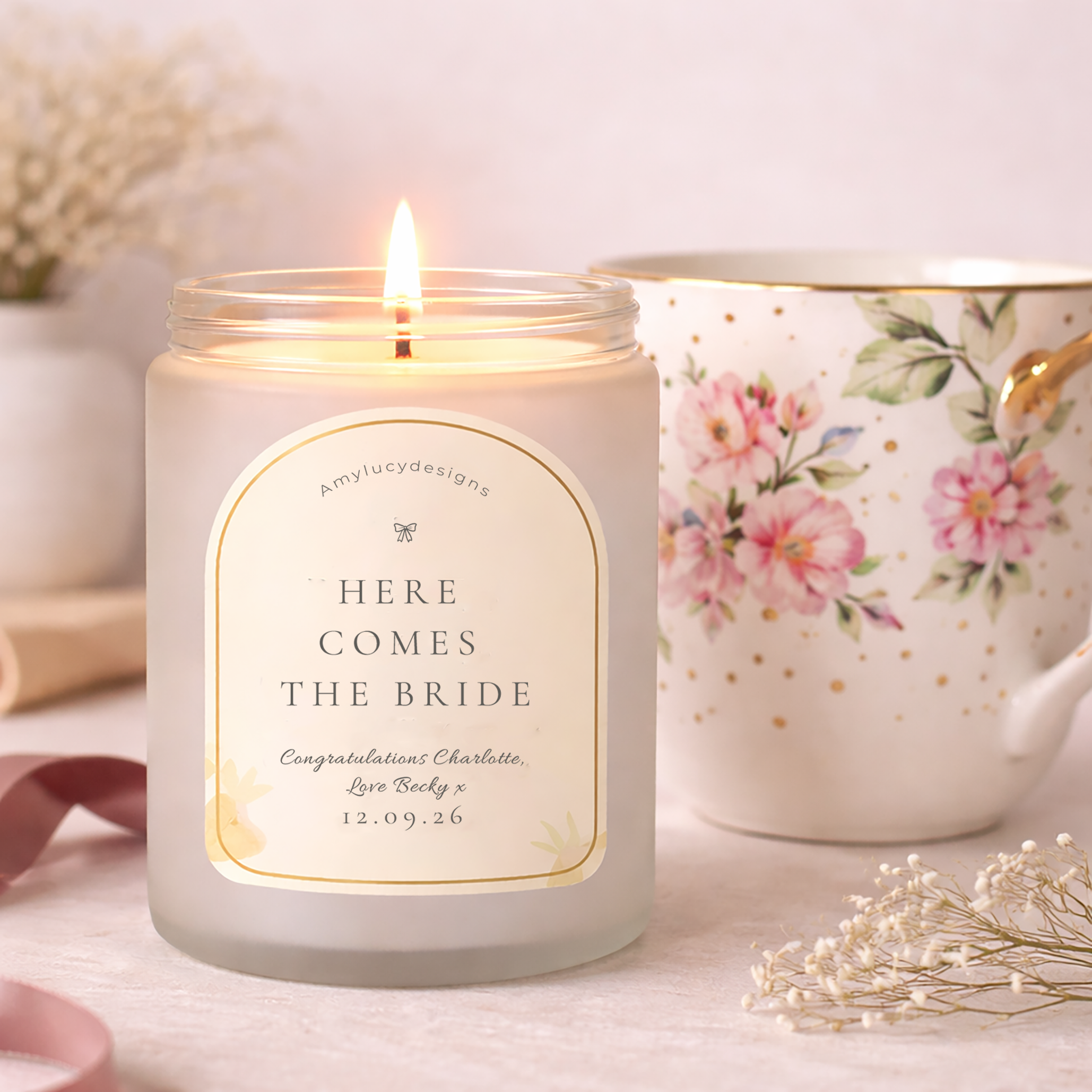 Candle with 'Here Comes the Bride' message in a decorative jar on a table with flowers and teacup.