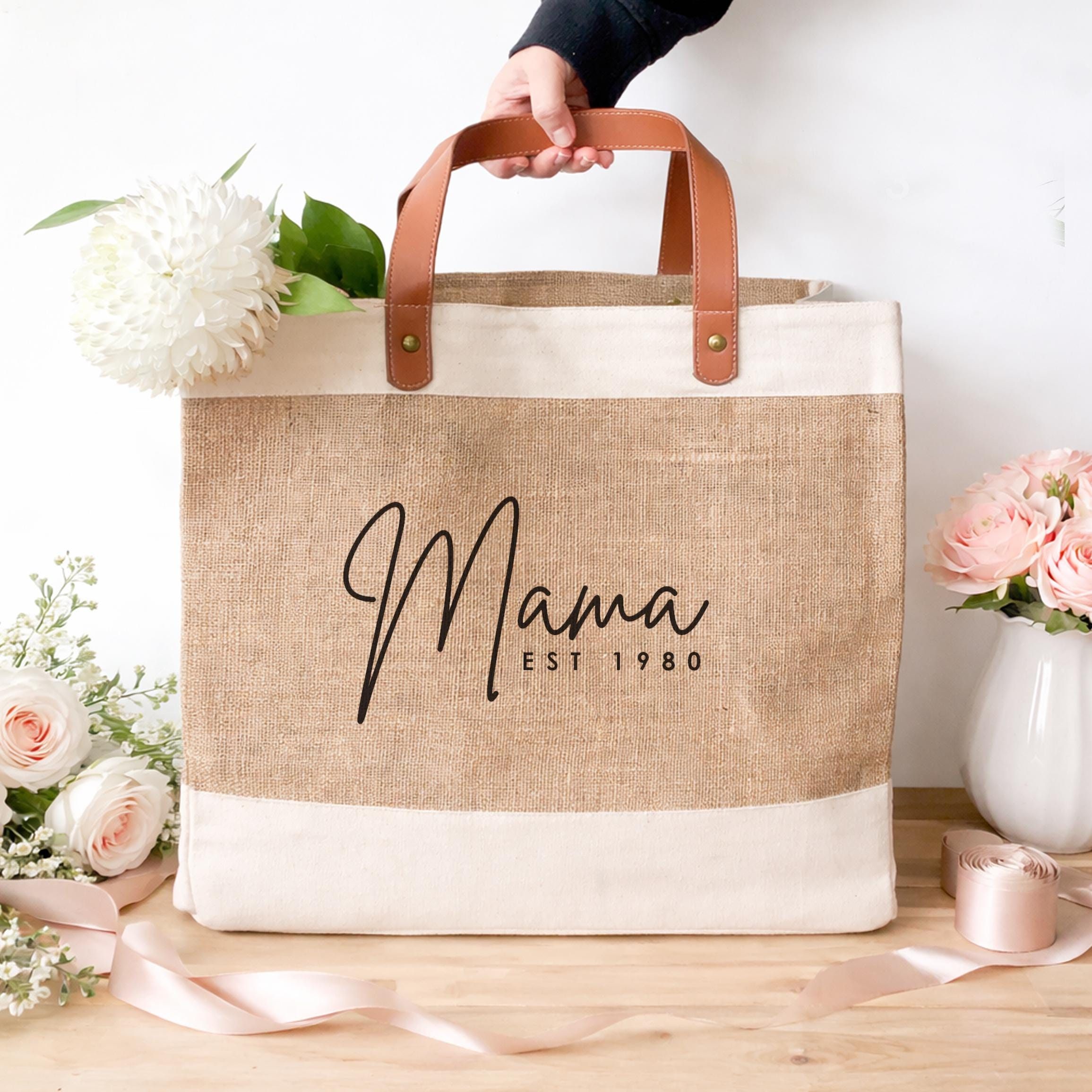 Jute bag with 'Mama Est. 1980' text held by a hand on a wooden surface with flowers.