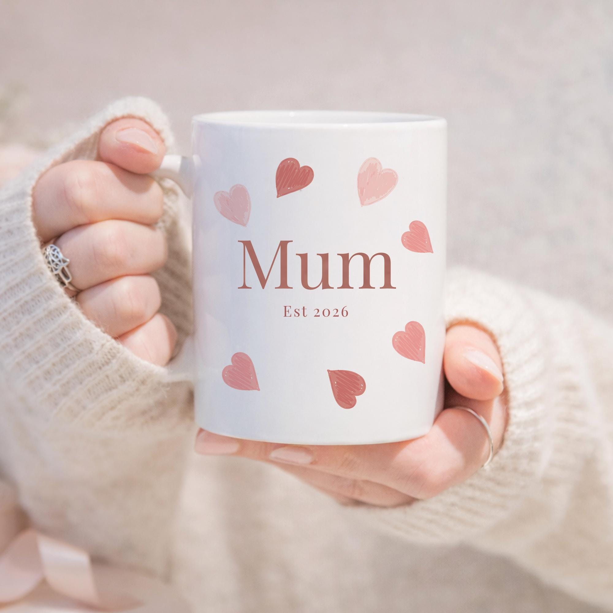 White mug with 'Mum Est 2026' text and pink hearts held by a person wearing a white sweater.