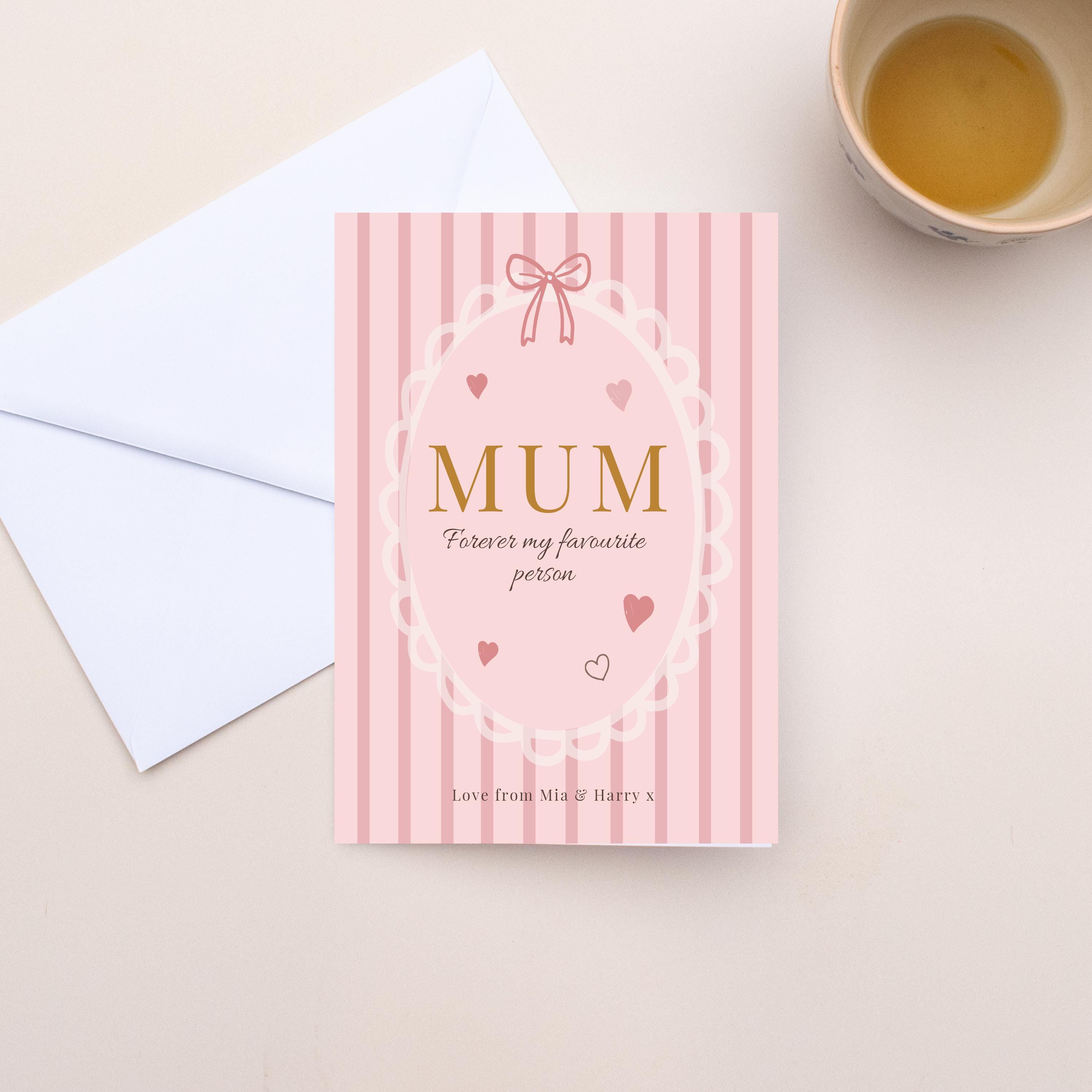 Pink card with 'MUM Forever my favourite person' text on a beige surface with a white envelope and cup.