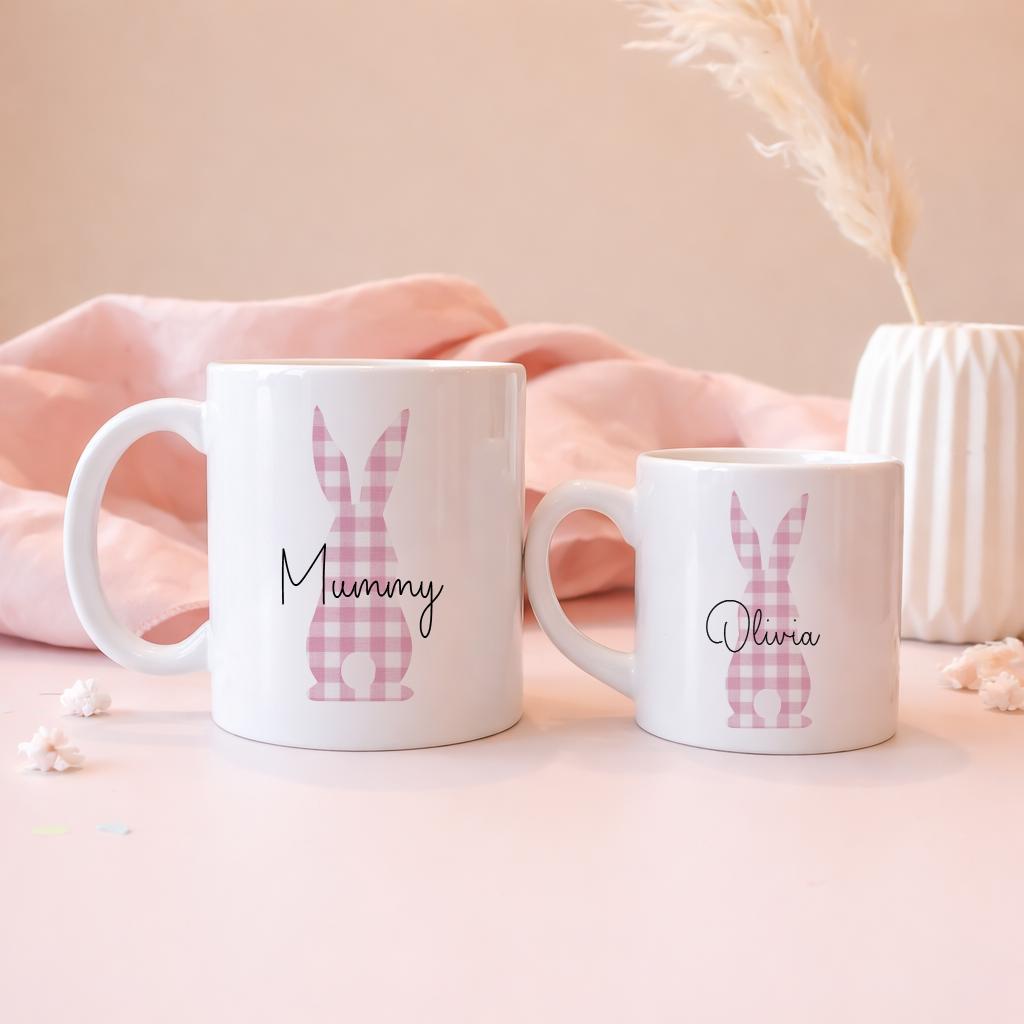 Two white mugs with pink bunny designs and names on a soft pink background