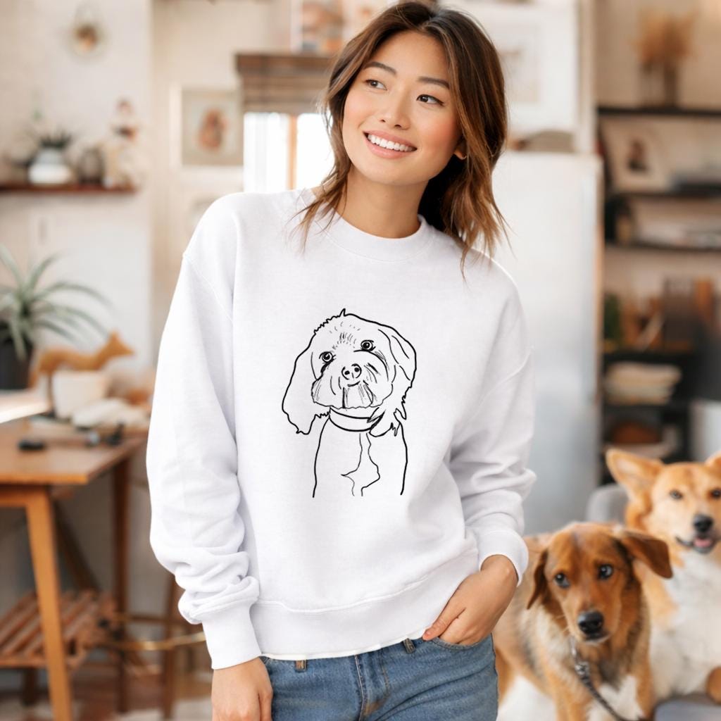 Woman wearing a white sweatshirt with a dog illustration, standing in a home setting with two dogs.